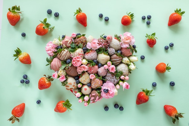 Caixa em forma de coração com morangos cobertos de chocolate artesanais com diferentes coberturas e flores como presente no dia dos namorados em fundo verde