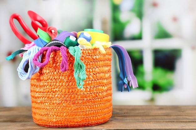 Caixa laranja redonda cheia de fios multicoloridos e tudo o que é necessário para costurar em uma mesa de madeira marrom em frente à janela