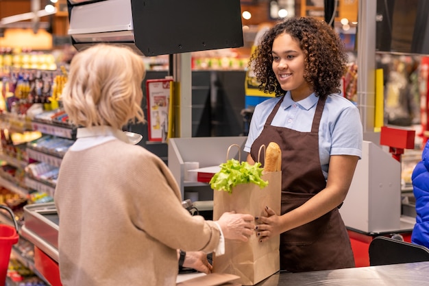 Caixa muito jovem dando uma bolsa de papel feminina madura com pão e mantimentos frescos, enquanto as duas estavam perto da caixa registradora no supermercado