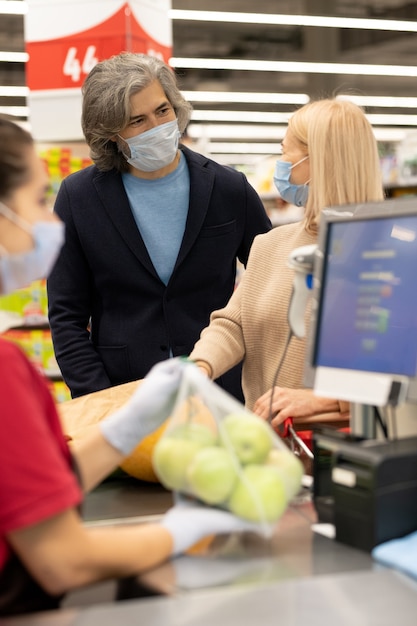 Caixa muito jovem dando uma bolsa de papel feminina madura com pão e mantimentos frescos, enquanto as duas estavam perto da caixa registradora no supermercado