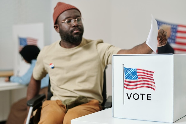 Foto caixa para boletins de voto na mesa e homem negro com deficiência fazendo sua escolha