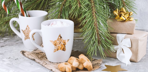 Caixas de presente, caneca com bebida decorada com marshmallow e biscoitos em forma de estrela perto de galhos de árvores de natal sempre-verdes