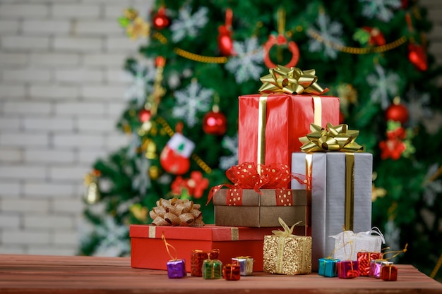 Caixas de presente coloridas pequenas e grandes com gravata borboleta de fita brilhante colocada no canto da mesa de madeira em frente totalmente decorativo lindo pinheiro de véspera de Natal e parede de tijolos no fundo desfocado.