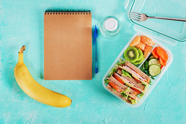 Caja de almuerzo escolar con sándwich, verduras, agua y frutas en la mesa.