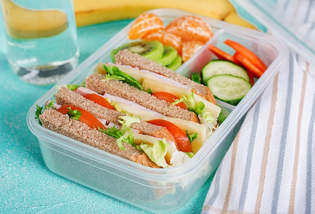 Caja de almuerzo escolar con sándwich, verduras, agua y frutas en la mesa.