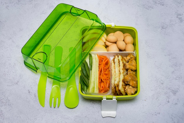 Caja de almuerzo en verde con cubiertos Merienda con pollo pepinos zanahorias hojas de espinaca Rebanada de manzana y galletas para el postre Alimentación saludable Espacio de copia