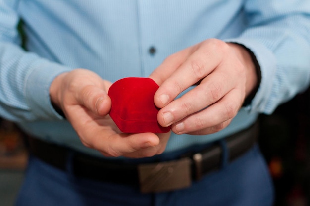 Una caja de anillo de terciopelo rojo cerrada en manos de un hombre con una camisa azul