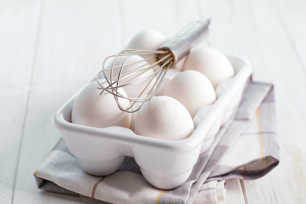 Caja de cerámica con huevos de granja orgánicos naturales y batidor de metal sobre una mesa de cocina de madera.