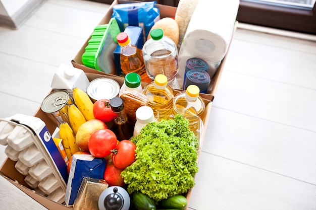 La caja ecológica de cartón completa con productos de la tienda de comestibles en el piso de la casa cerca de la puerta