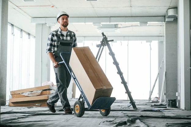Con una caja grande que transporta el material Trabajador de la construcción en uniforme en una habitación vacía sin terminar