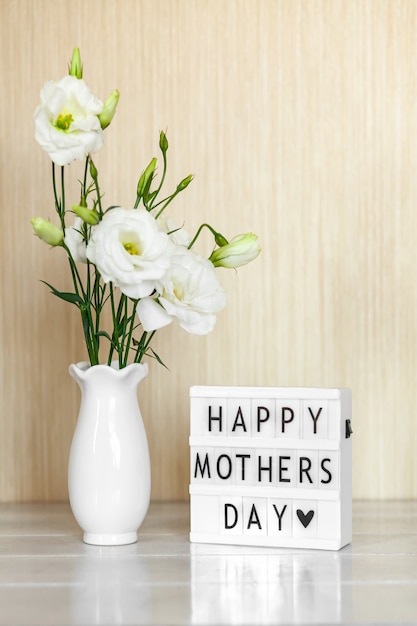 Caja de luz con letras Feliz día de la madre, flores blancas Eustoma o Lisianthus en florero sobre mesa de madera.