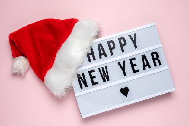 Caja de luz con texto FELIZ AÑO NUEVO y sombrero de Santa Claus sobre fondo rosa pastel. Concepto mínimo de Navidad o año nuevo.