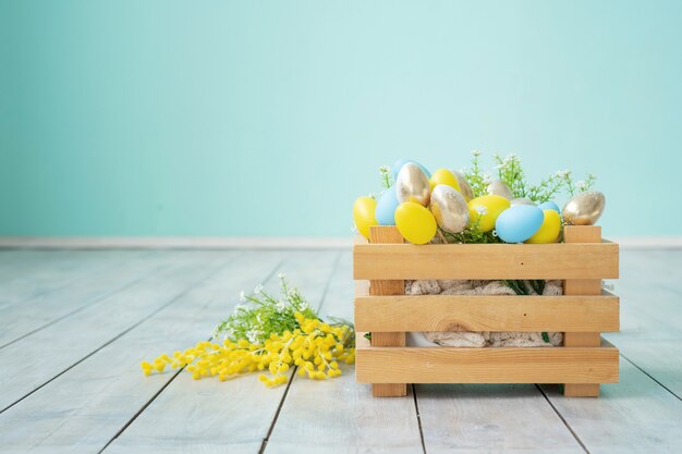 Caja de madera con huevos de Pascua azules, amarillos y dorados contra una pared azul