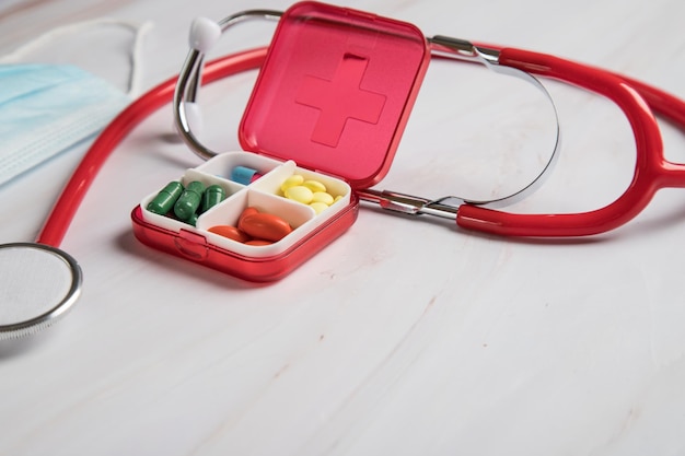 Una caja con pastillas de colores, un estetoscopio y una máscara médica sobre una mesa de luz