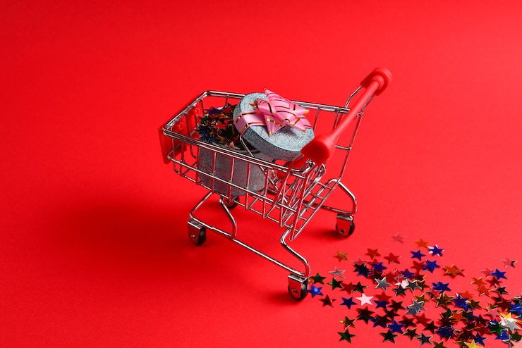 Foto caja de regalo azul con lazo rosa en un carrito de compras y confeti sobre un fondo rojo.