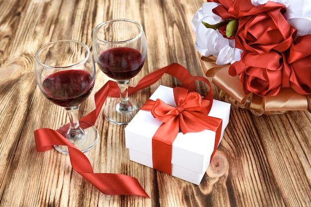 Caja de regalo blanca con lazo de cinta de raso rojo, dos copas de vino y ramo de flores rosas rojas y blancas artificiales sobre fondo de madera.