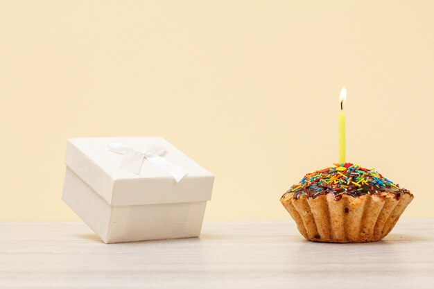 Caja de regalo blanca y sabroso muffin de cumpleaños con glaseado de chocolate y caramelo, decorado con velas festivas encendidas sobre fondo beige. Feliz cumpleaños concepto mínimo.