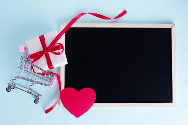 Caja de regalo en carros de compras con corazón de papel rojo y pizarra en blanco