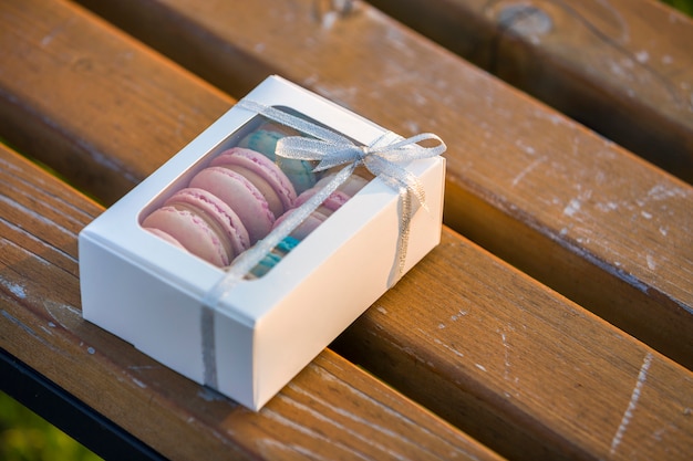 Caja de regalo de cartón blanco con coloridas galletas macaron hechas a mano en un banco de madera al aire libre.