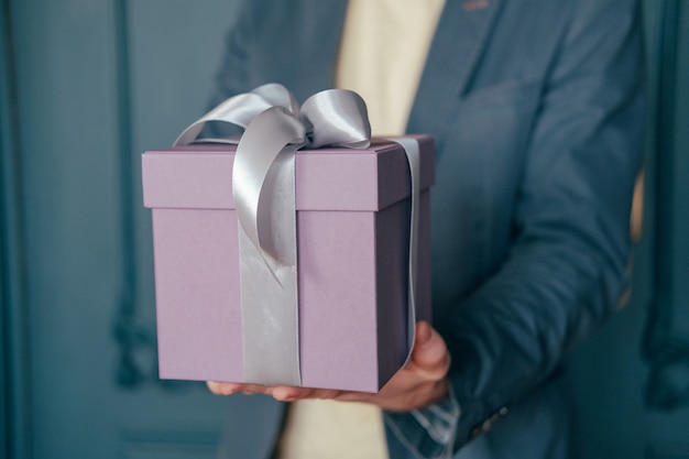 Caja de regalo con cinta de plata gris en las manos de un hombre elegante sobre un fondo azul