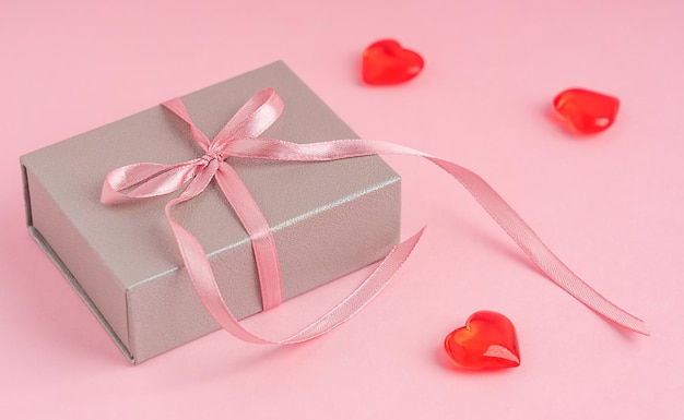 Una caja de regalo de color plateado con lazo hecho de cinta textil sobre fondo rosa con decoración de corazón rojo