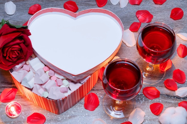 Caja de regalo del Día de San Valentín feliz en forma de corazón y dos copas de pétalos de rosa de vino tinto se encuentran