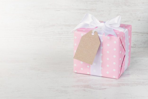 Caja de regalo envuelta en papel punteado rosa y lazo de satén atado sobre madera blanca.