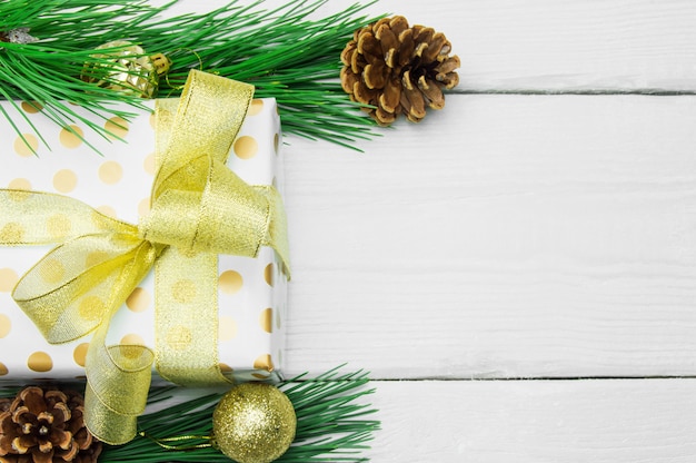 Caja de regalo con lazo de cinta dorada y rama de árbol de Navidad con cono en superficie vintage de madera blanca