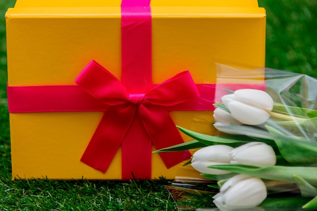 Caja de regalo con lazo rosa y tulipanes en césped de hierba verde