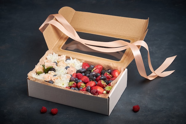 Caja de regalo llena de flores blancas y rosadas y frutas maduras sobre un fondo oscuro