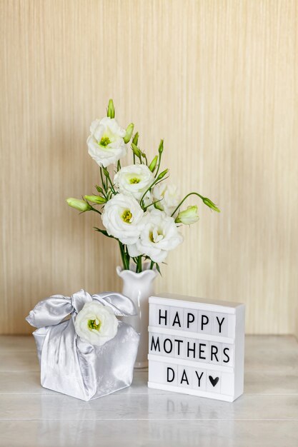 Caja de regalo de moda envuelta en tela de seda en técnica Furoshiki, caja de luz con letras Feliz Día de la Madre, flores blancas Eustoma o Lisianthus en florero sobre mesa de madera. Concepto de vida cero residuos.