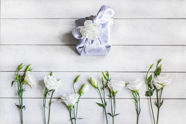 Caja de regalo de moda envuelta en tela de seda en la técnica Furoshiki flores blancas Eustoma o Lisianthus yacen en una fila en la parte inferior sobre fondo de madera clara vista superior plano Lay Zero Waste Life Concept