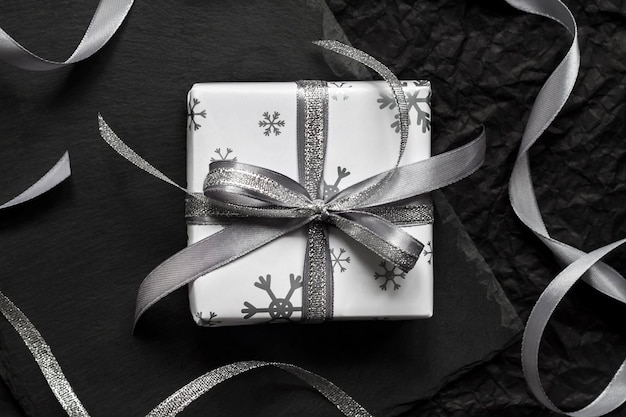 Caja de regalo de Navidad envuelta en un papel blanco con copos de nieve y atada con una cinta sobre fondo oscuro. Endecha plana, vista superior