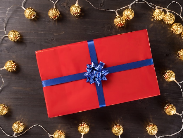 Caja de regalo de Navidad roja con cajas de regalo más pequeñas sobre un piso de madera oscura rodeado de luces doradas. Ambiente feliz