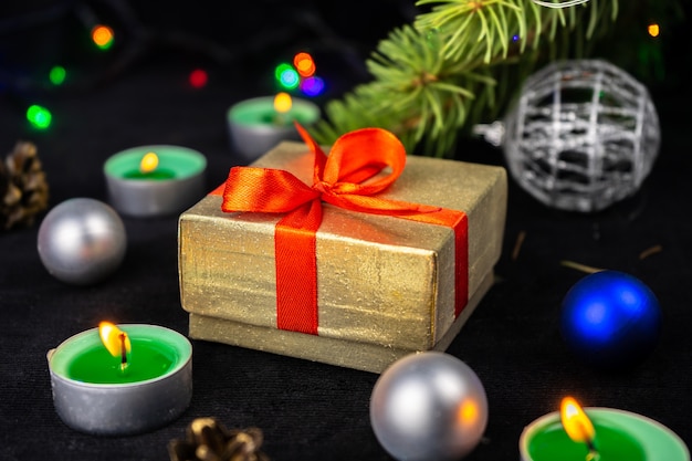 Caja de regalo de oro presente con cinta roja junto al árbol de Navidad, velas y decoración navideña.