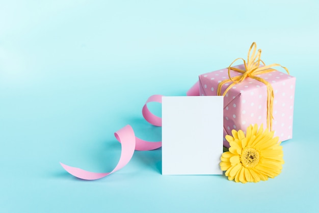 Caja de regalo punteada rosa, flor de gerbera amarilla y tarjeta vacía sobre azul.