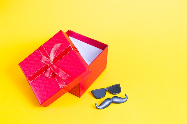 Caja de regalo roja y bigote y gafas en miniatura sobre un fondo amarillo, espacio de copia