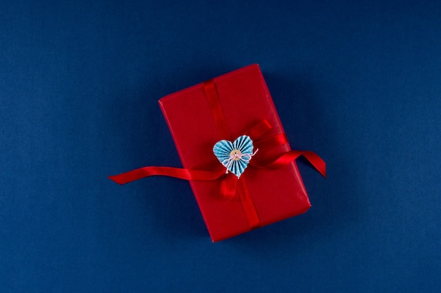 Caja de regalo roja con corazón y lazo sobre fondo de color azul clásico 2020. Concepto de empaquetado del día de San Valentín 14 de febrero. Endecha plana, espacio de copia, vista superior.