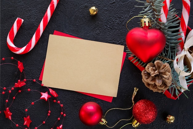 Caja de regalo roja con un juguete navideño en forma de corazón, ramas de abeto, dulces navideños, guirnalda y tarjeta de texto de felicitación sobre el fondo negro