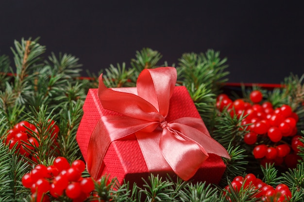 Caja de regalo roja presente con lazo rosa satinado, inmerso en las agujas de un árbol de Navidad decorado con bayas rojas.