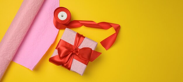 Caja de regalo rosa envuelta en cinta de seda roja sobre fondo amarillo, espacio de copia