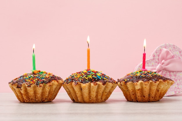 Caja de regalo y sabrosos muffins de cumpleaños con glaseado de chocolate y caramelo, decorados con velas festivas encendidas sobre fondo de madera y rosa. Concepto de feliz cumpleaños.