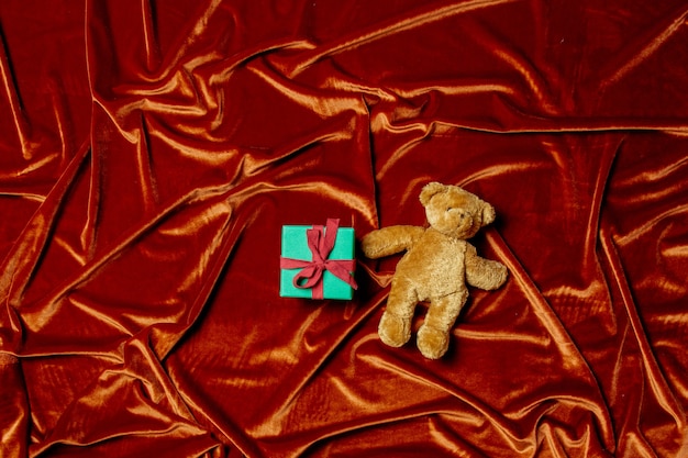 Caja de regalo de vacaciones con osito de peluche sobre fondo de tela de color rojo carmesí.