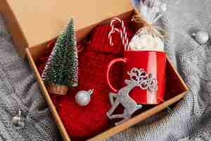 Foto una caja con regalos para navidad y año nuevo una taza roja con malvaviscos guantes de punto juguetes de navidad y un pequeño árbol de navidad sobre un fondo de punto gris