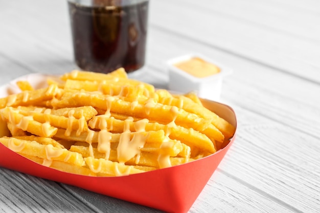 Caja roja de papel llena de sabrosas papas fritas con salsa de queso sobre fondo blanco de madera