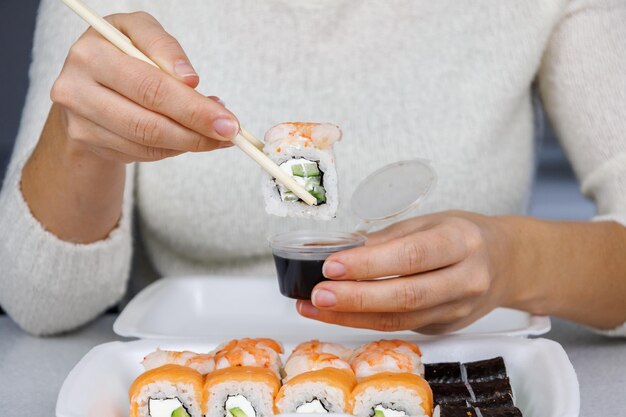 Una caja de rollos de Filadelfia sobre la mesa en la cocina Sushi de entrega rápida en un recipiente blanco La niña come con palillos Decoración festiva Concepto de Navidad