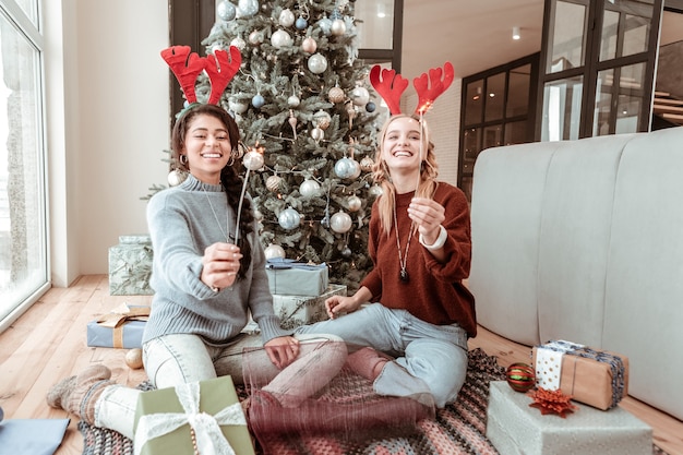 Cajas para amigos. Alegres niñas felices sentadas en el piso de la sala de estar y llevando bengalas mientras están de fiesta. Concepto de navidad