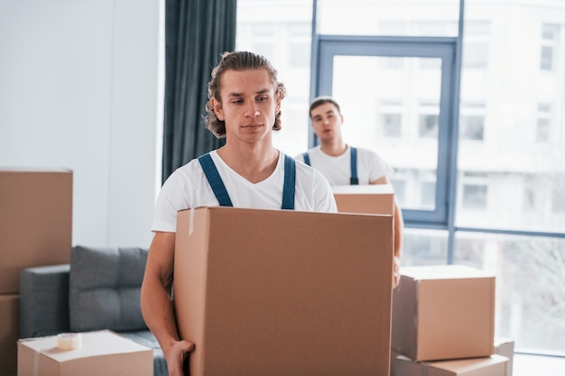 Con cajas en las manos Dos jóvenes motores en uniforme azul trabajando en el interior de la habitación