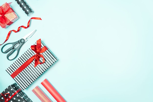 Cajas de regalo envueltas en papel rojo y a rayas en blanco y negro con materiales de embalaje sobre un fondo azul. Preparación de regalos de Navidad.
