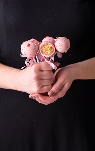 Cake pops en manos de las mujeres sobre un fondo negro. Postre en crema de chocolate rosa con polvo y lazo.